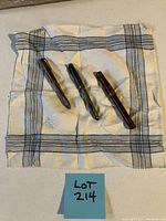 Photo showing three vintage fountain pens laid out on a plaid patterned handkerchief with a lot label.