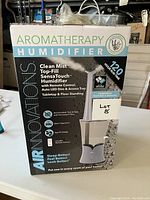 Front view of the unopened box showing the product name and features such as clean mist, top-fill, SensaTouch, remote control, auto LED dim, aroma tray, and 20 hours run time.