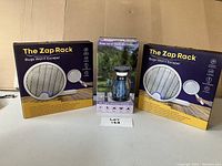 Image showing two boxed 'The Zap Rack' handheld electric bug swatter zappers and one boxed solar-powered UV insect zapper lantern.