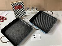 Two square cooking pans, four small glass spice jars, and a cookbook on a white table.