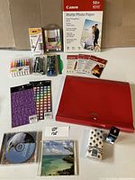 Full lot view showing all items arranged on a surface: Canon photo paper box, colored felt pens, styluses, storage file, alphabet stickers, adhesive tape, CDs, photo slides