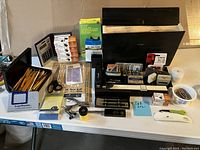 Photo showing a black calculator, a container full of wood pencils, black stapler with extra staples, a black tape dispenser, white shoehorn with Naturalizer branding, and other small office supplies scattered on the table.