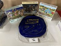 View of three boxed puzzles and a blue puzzle assembly system mat in original packaging, set on a surface with a lot number card.