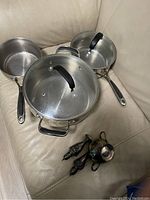 Three stainless steel cooking pots arranged on beige leather surface. Two pots contain glass lids with black handles, one pot without lid visible in corner. Black handles on pots are heat resistant.