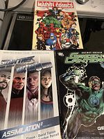Three graphic novels stacked showing front covers: Marvel Comics Origins with many Marvel heroes, Star Trek Voyager Assimilation² Volume 2 featuring crew members and Doctor Who, and DC Green Lantern Secret Origin with a glowing Green Lantern on the cover.