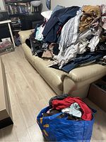 Large pile of assorted men’s and women’s clothing on a beige leather couch including jeans, pants, shirts, and sweatshirts.