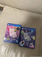 Two PS4 game cases: Just Dance 2020 and Just Dance 2022, resting on a light colored fabric couch.