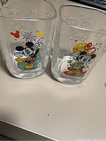 Photo of two clear glass Mickey Mouse cups with colorful designs and embossed Disney logos.