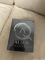 Sealed Alien Quadrilogy DVD box set placed on a beige leather couch. Front of the box shows the Alien logo and text 'Alien Quadrilogy'.