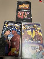 Photo showing three WrestleMania VHS tapes. One tape is WrestleMania XII from 1996 showing wrestlers Shawn Michaels, Bret Hart, Diesel, and The Undertaker on the cover. Two WrestleMania VI tapes also shown: one with Hulk Hogan and The Ultimate Warrior face-to-face on the cover, the other featuring Sid Justice vs Hulk Hogan and Macho Flair Affair match on the cover.