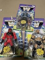 Photo showing three sealed WWE Masters of the WWE Universe figures, one blue, one red, one silver, in original packaging.