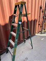 Full view of the green A frame ladder standing upright with yellow top cap beside a red container wall.