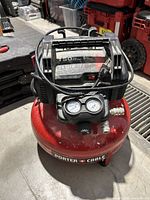 Red Porter Cable air compressor on concrete floor with power cord wrapped around handle and visible pressure gauges on front.