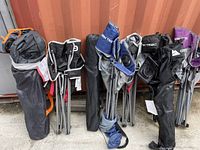 Lot of 8 foldable camping chairs in carry bags, standing upright against a metal container wall outdoors.