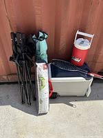 Camping lot including cooler, water jug, campfire grill basket, two foldable chairs, and a folded green camping item, all arranged against a red metal wall outdoors.