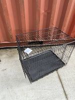 Top and side view of a black metal wire cage with a removable plastic tray, placed on concrete floor in front of a metal container wall.