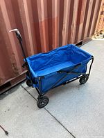 Blue collapsible wagon fully open showing handle, fabric interior, and wheels on concrete surface next to a shipping container.