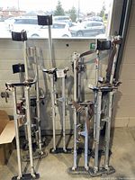 Several pairs of metal drywall stilts leaning against a wall, showing adjustable height mechanisms, foot platforms, padded calf supports, and straps. Used condition with dirt and wear visible.