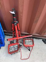 Red tripod work light with dual red metal light fixtures having black protective wire cages, placed on concrete surface near a container.