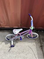 Full view of the pink kids bicycle with training wheels against a brown wall, showing frame, tires, seat, and handlebar.