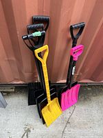 Five plastic shovels leaning against a container wall outdoors on concrete. Colors include black, yellow, and pink. Handles have ergonomic grips; blade styles vary including flat and rake-like.