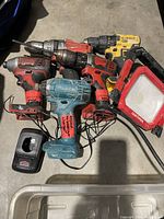 Power tools grouped on floor showing multiple cordless drills from Milwaukee, DeWalt, Makita, and a Craftsman work light, with visible wear, labels showing test status, missing batteries and chargers.