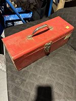 Closed red metal toolbox with a latch and handle, sitting on a black surface