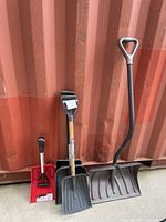 Four snow shovels leaning against a corrugated metal backdrop, showing variety in size and handle types.