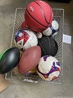 Top-down view of a wire basket containing various sports balls showing basketballs, soccer balls, footballs, and baseballs in assorted colors and brands.