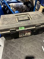 Closed black plastic tool box showing wear, dust and a label on the front.