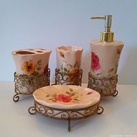 Front view of four ceramic bathroom accessories in decorative metal holders, showing floral prints and gold pump dispenser