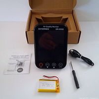 Photo of Goyerrnes GR-533G air quality monitor with touchscreen, rechargeable battery, USB charging cable, small screwdriver and user manual on white surface. Device screen is off reflecting surroundings.