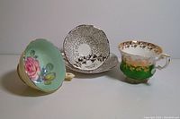 Three vintage teacups and saucers: green gold Queen Anne, black and white Royal Albert, and floral Paragon with pink rose interior.