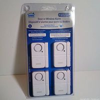 Photo of the unopened original blue and white packaging containing four white Sabre door/window alarm sensors. Package text highlights features such as 120dB siren, low battery test button, 4-pack, and easy installation.