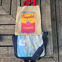 Two bags placed on wooden surface: beige Campbell's soup tote with red handles and colorful design; blue/black messenger bag with NYC subway map print
