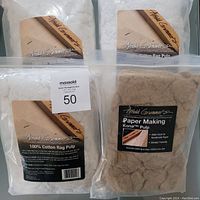 Four packages of Arnold Grummer's paper making pulp, three white cotton rag pulp and one brown Kona tm pulp, shown in sealed plastic bags with labels mostly visible.