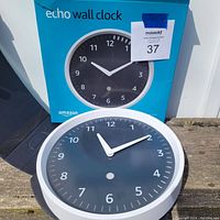 Amazon Echo Wall Clock with black face, white frame, white numbers and hands, placed in front of its original blue box packaging.
