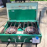 Full view of Coleman green dual burner propane camping stove with lid open showing two rusted burners and metal grate on wooden surface outdoors.