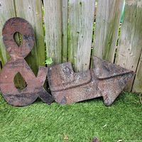 Galvanized steel ampersand and arrow signs placed on grass in front of a wooden fence, showing rust and weathering on surface.