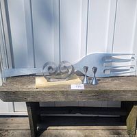 Full view of giant aluminum fork with long handle and three tines, pair of Umbra taper candle holders, and vintage double coil book holder arranged on a wooden bench.