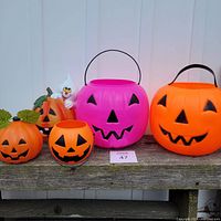Lot showing five pumpkin and ghost themed Halloween decorations and buckets on wooden surface against wall background. Includes two large pumpkin buckets and 3 small pumpkin/ghost decorations.