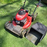 Red Troy-Bilt self-propelled lawnmower on grass next to a black detachable grass collection bag showing wheels and handle