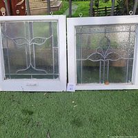 Two leaded glass window panels set in white frames, on grass outdoors with hooks on top visible, showing both panels side by side.