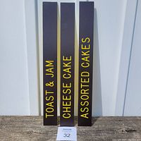 Three brown plastic menu slider signs with yellow lettering standing upright on a wooden surface against a white paneled background. Signs read Toast & Jam, Cheese Cake, and Assorted Cakes.