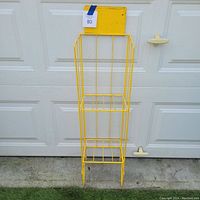 Full front view of the yellow metal wire magazine display rack standing on concrete with garage door background.