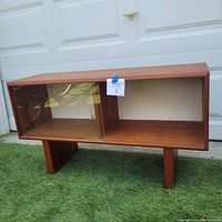 Front view of small teak veneer Mid-Century Modern credenza cabinet with one glass sliding door and open compartment.