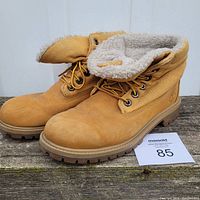 Pair of tan Timberland boots with shearling lining visible on the folded collars and rugged soles, set on wooden surface against white background.