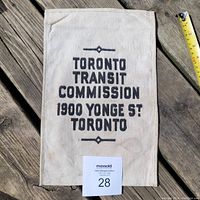 Full view of off-white canvas coin token bag with printed black text 'TORONTO TRANSIT COMMISSION 1900 YONGE ST TORONTO'. Bag laid flat on wood deck next to tape measure showing length.