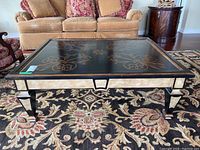 Front view of antique style coffee table showing detailed gold Italian floral designs on black tabletop and tapered legs with faux marble finish.