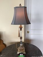 Full view of antique style table lamp on a black marble table, showcasing the silk gray shade and metal base with clear glass column.